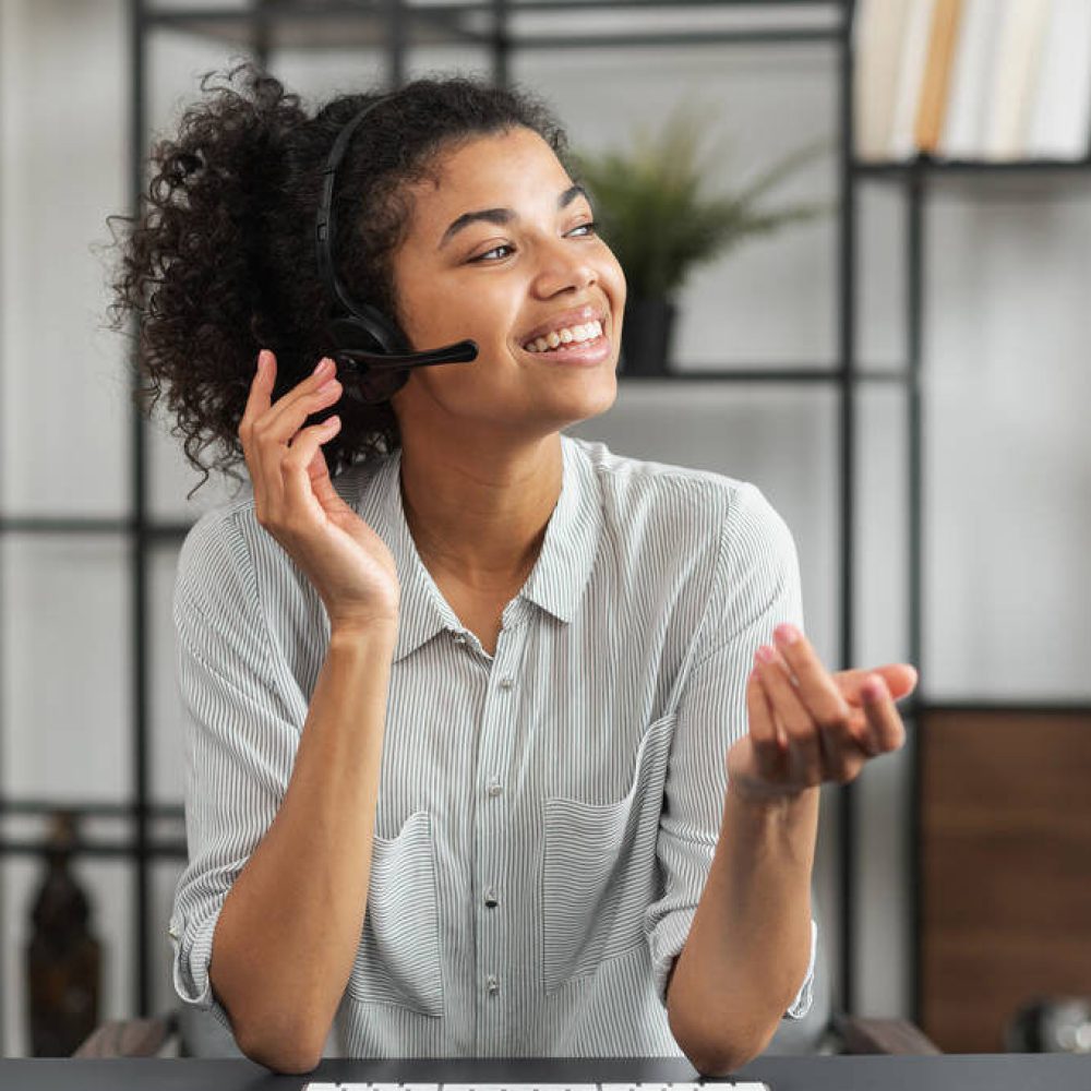 Young smiley African American female customer service agent in a headset, touching microphone, talking and looking away, enjoying her work, and feeling happy to help clients and satisfy their needs